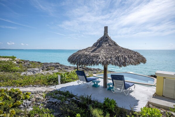 Un espace piscine agrémenté d’un parasol en chaume, de chaises longues et offrant une vue sur l’océan.