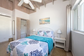 Une chambre à coucher avec un lit, des tables de chevet, un ventilateur de plafond, un tableau et une fenêtre.