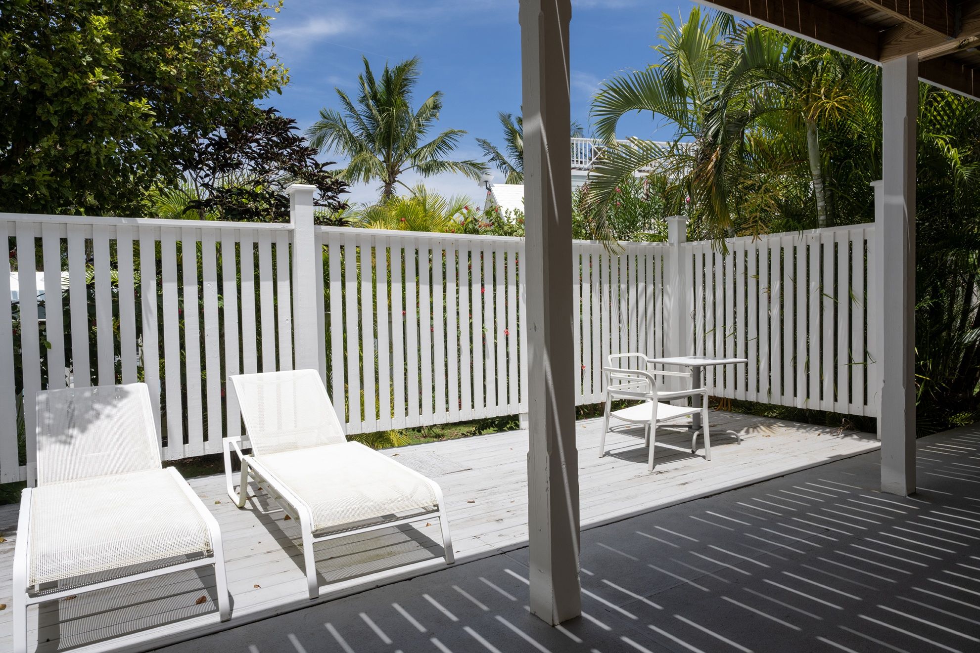 A covered outdoor area with a white fence, two white lounge chairs, and a small table.