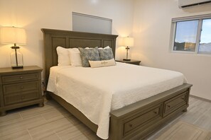 A bedroom with a large bed, two bedside tables with lamps, a window, and a wall-mounted air conditioner. - Emerald Cottage - Brand New Beach Cottage With Pool, Walk To La 1 Bedroom Home (North Palmetto Point)