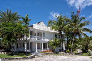 Un bâtiment blanc à deux étages, doté d’un balcon, entouré de palmiers et surplombé d’un ciel bleu azur.