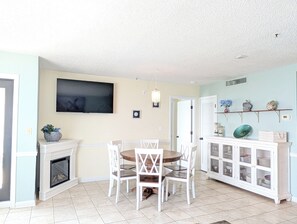 A dining area with a white table and chairs, a fireplace, a television mounted on the wall, and a cabinet with glass doors. - Ocean Front 2 BR | Myrtle Beach Resort - RT1902 (Myrtle Beach)