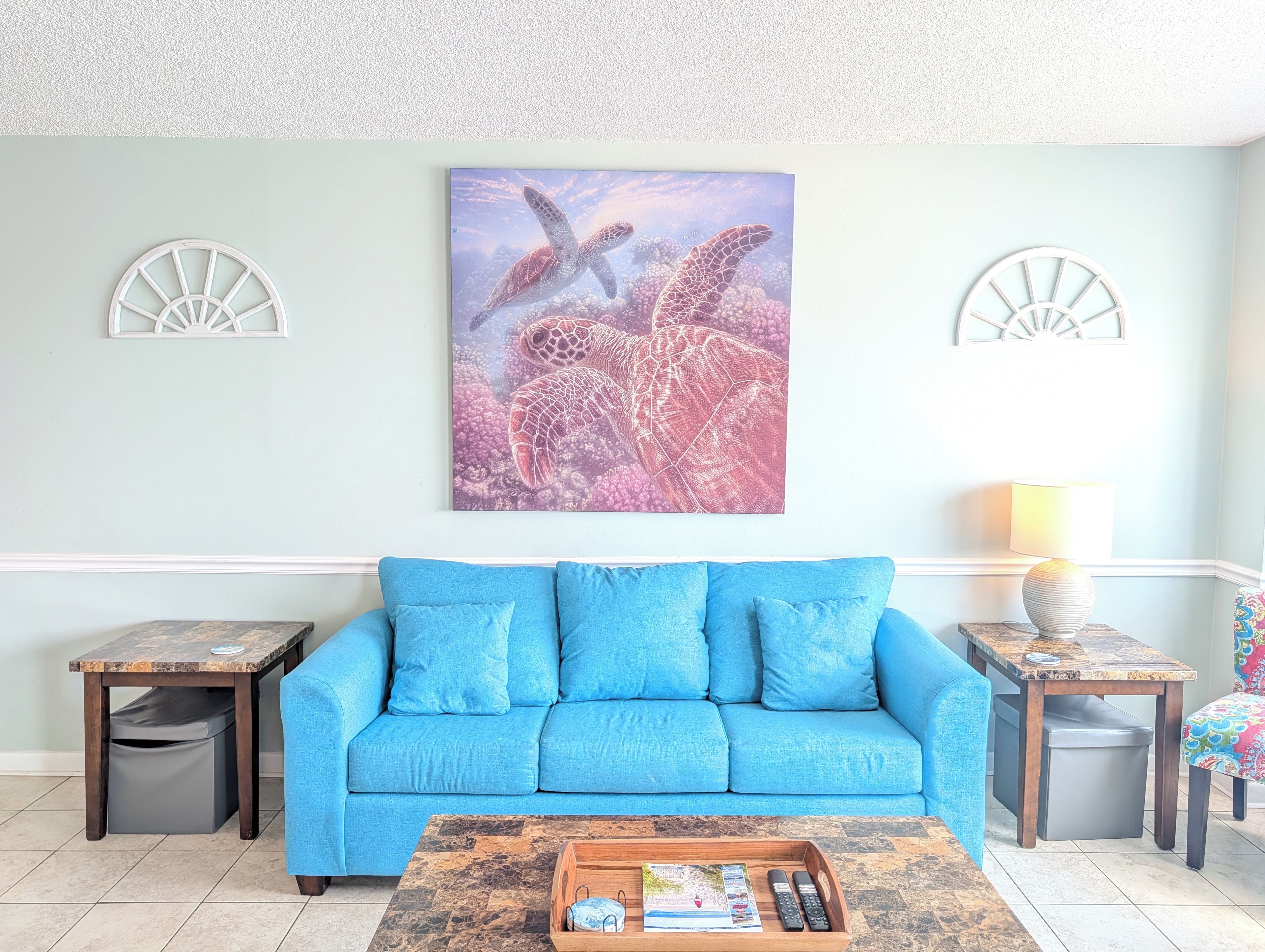 A living room with a blue sofa, a coffee table with remote controls, and a wall-mounted artwork of a sea turtle.
