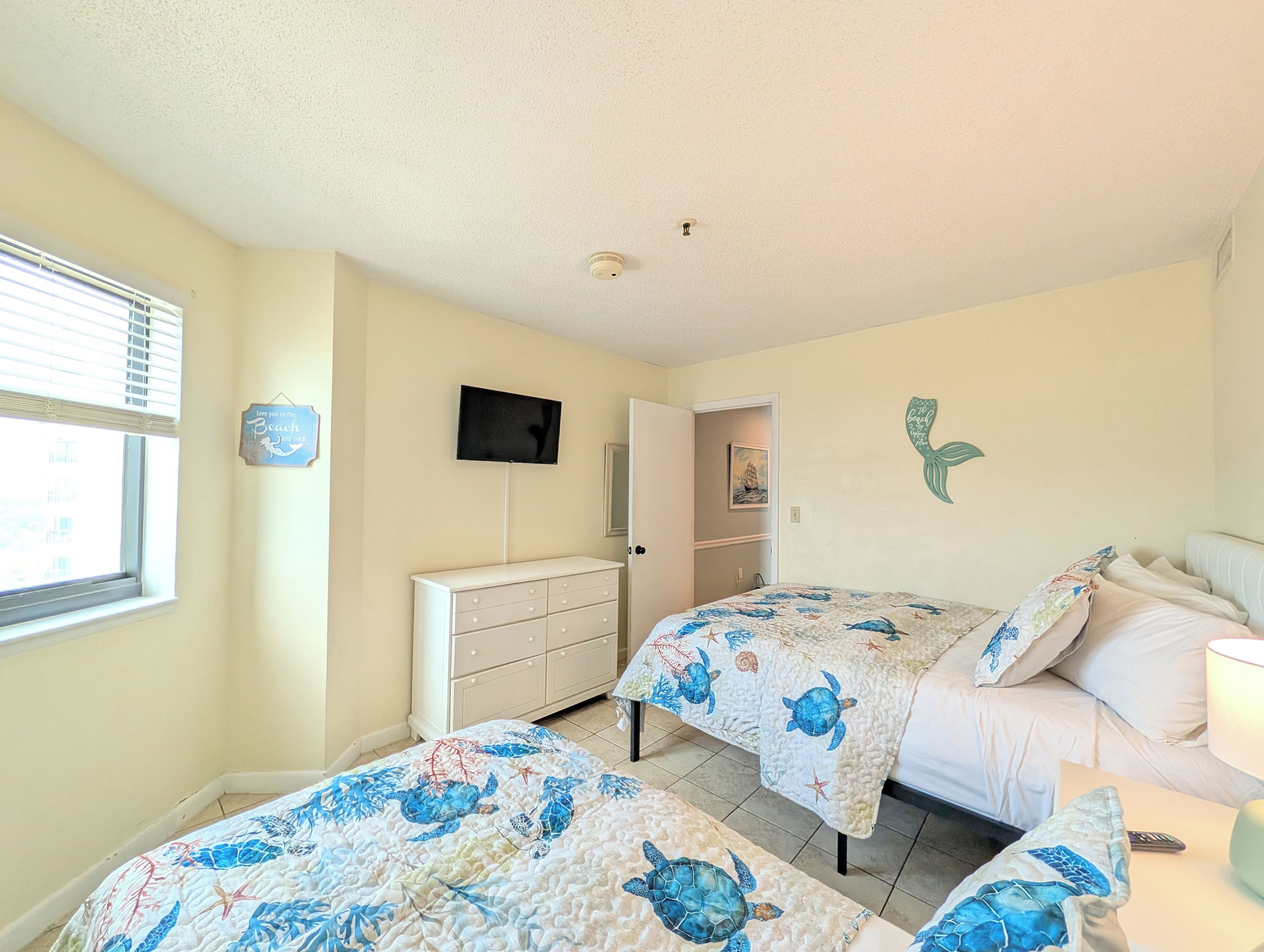 A bedroom with two beds, a dresser, a television, and a window with blinds.