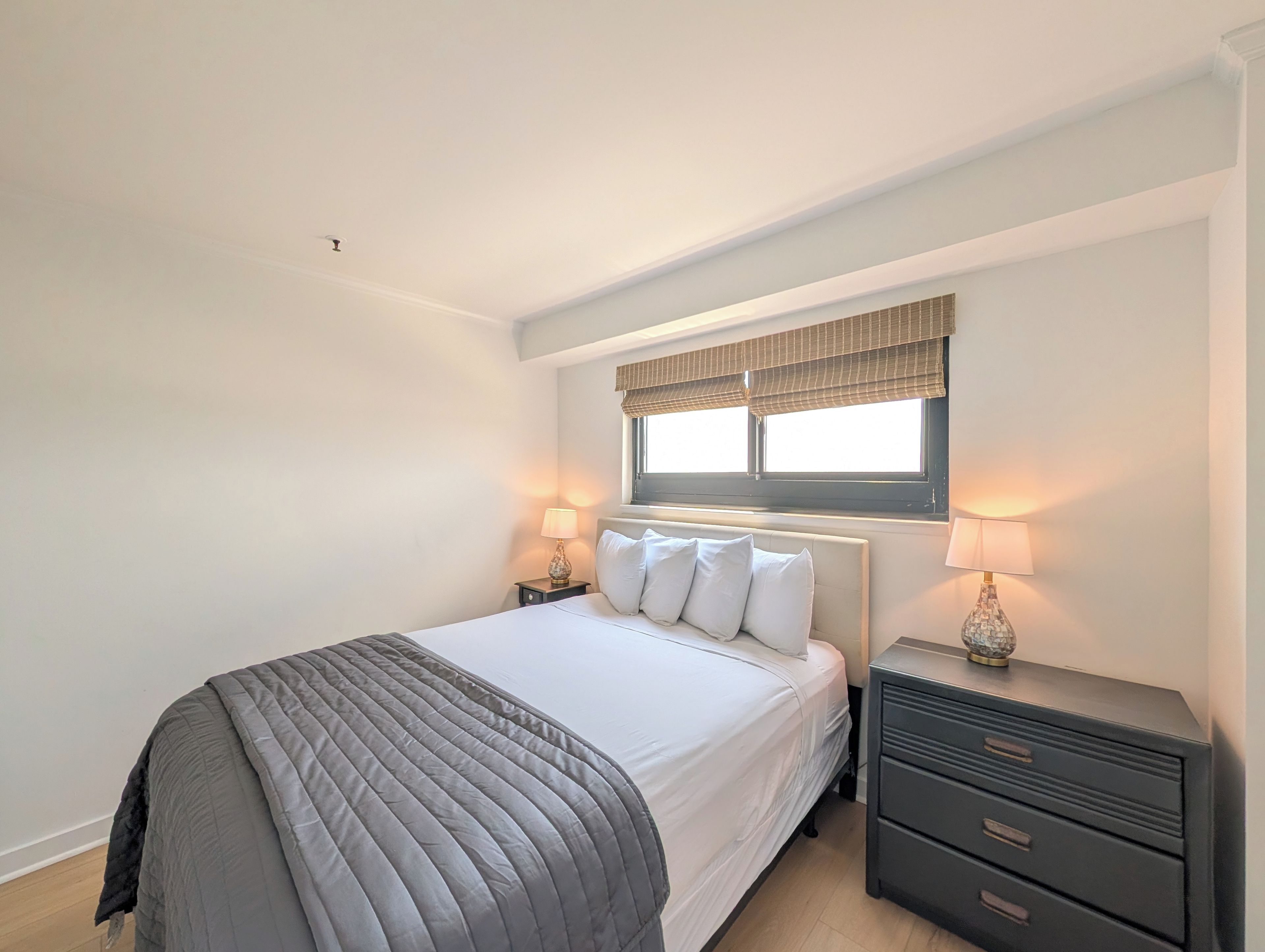 Une chambre à coucher avec un lit, des tables de chevet, une fenêtre avec des stores et une petite table de nuit.