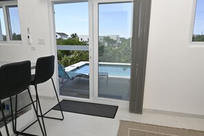Una habitación con puerta de vidrio corrediza que da a una zona de piscina, una pequeña mesa con sillas y vistas del vecindario circundante.
