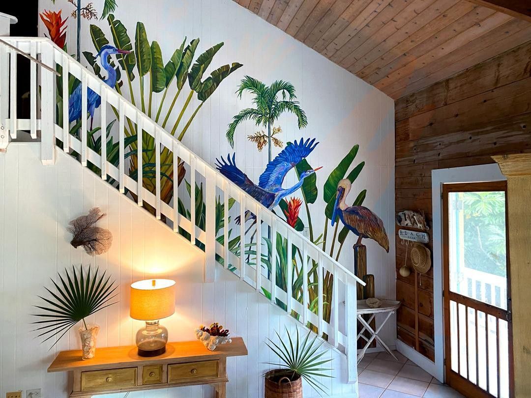 Escadaria com um mural de aves e plantas tropicais, corrimão de madeira e luminária sobre um console.