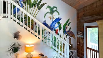 Escadaria com um mural de aves e plantas tropicais, corrimão de madeira e luminária sobre um console.