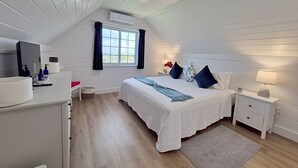 A bedroom with a sloped ceiling, a bed with a white duvet, a bedside table with a lamp, and a window with curtains. - Eagle Ray - Brand New Beach Cottage W/views, Walk To Restaurant/ 2 Bedroom Home (North Palmetto Point)