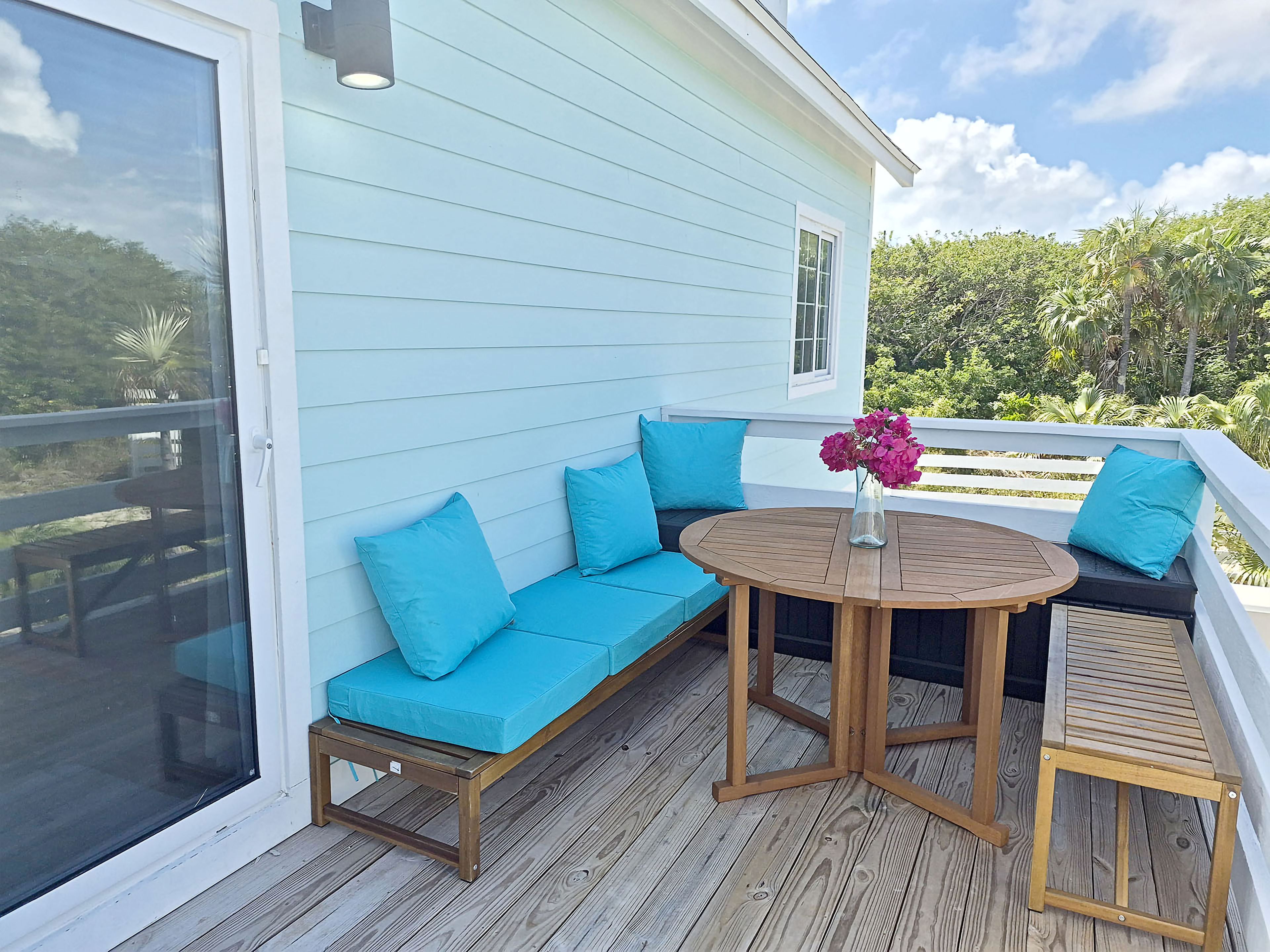 A covered outdoor seating area with a wooden table and benches, a glass door, and a view of greenery.