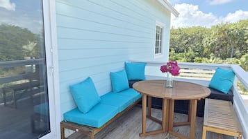 A covered outdoor seating area with a wooden table and benches, a glass door, and a view of greenery.