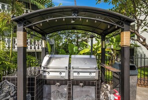 Een barbecue-opstelling met twee barbecues onder een overdekte constructie, omringd door weelderige begroeiing.