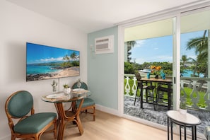 A room with a glass table, two chairs, a painting of a beach, and a sliding glass door leading to an outdoor area with a view of the ocean and a balcony. - Kona Islander Inn#251: Downtown Kona, Remodeled, Ocean view, Best Spot (Kailua Kona)