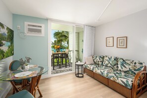 A room with a glass table, chairs, a sofa, and a view of a balcony with outdoor furniture. - Kona Islander Inn#251: Downtown Kona, Remodeled, Ocean view, Best Spot (Kailua Kona)