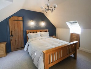 A bedroom with a sloped ceiling, a wooden bed, a nightstand, a dresser, and a chandelier. - Yr Hen Felin (Boncath)