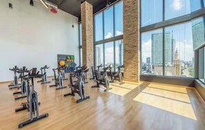 Ein Fitnessraum mit Ergometer, einem an der Wand befestigten Bildschirm und großen Fenstern mit Blick auf die Stadt.
