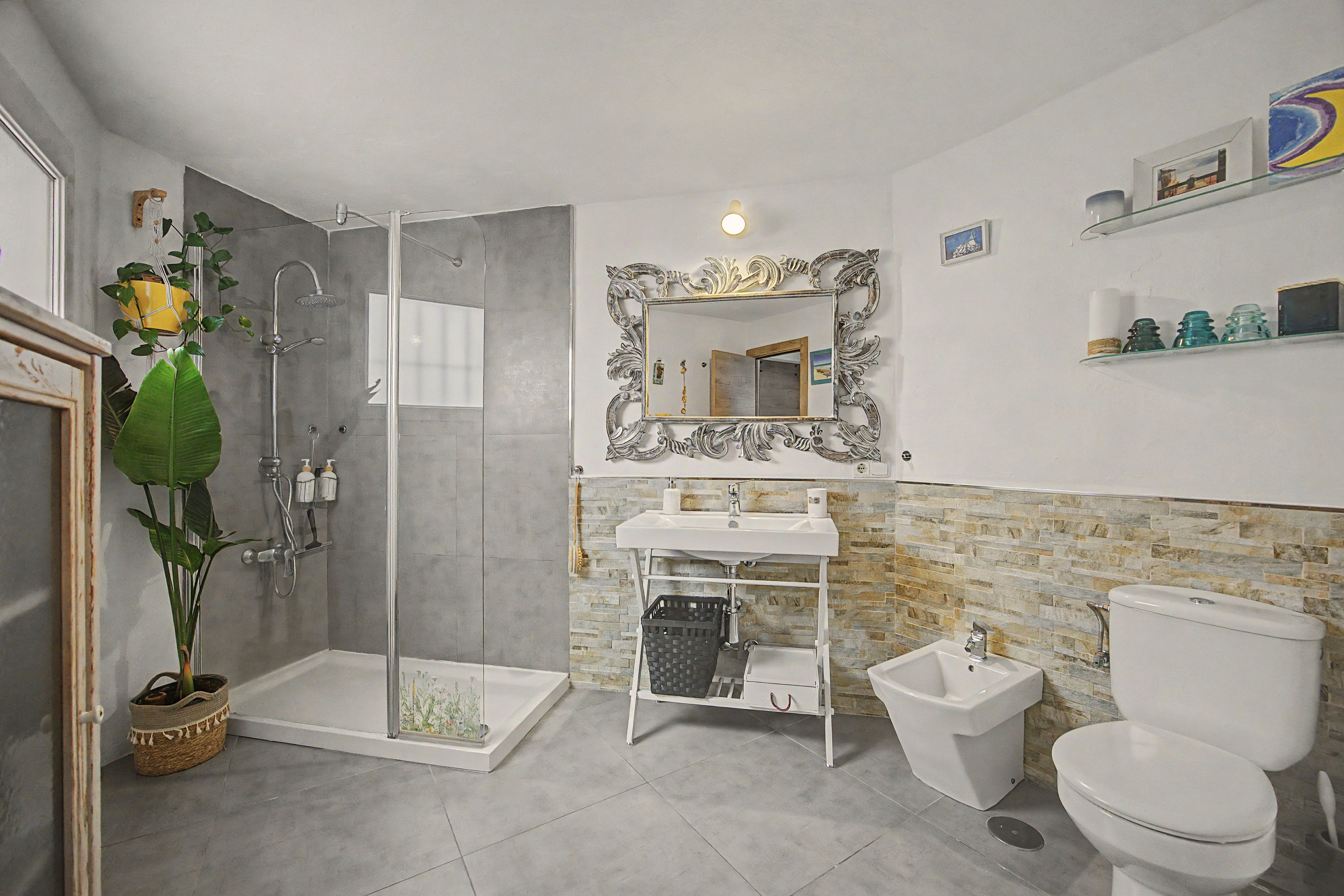 Ein modernes Badezimmer mit einer Duschkabine aus Glas, einem großen Spiegel mit aufwendigem Rahmen, einem weißen Waschbecken, einem Bidet und einer Toilette.