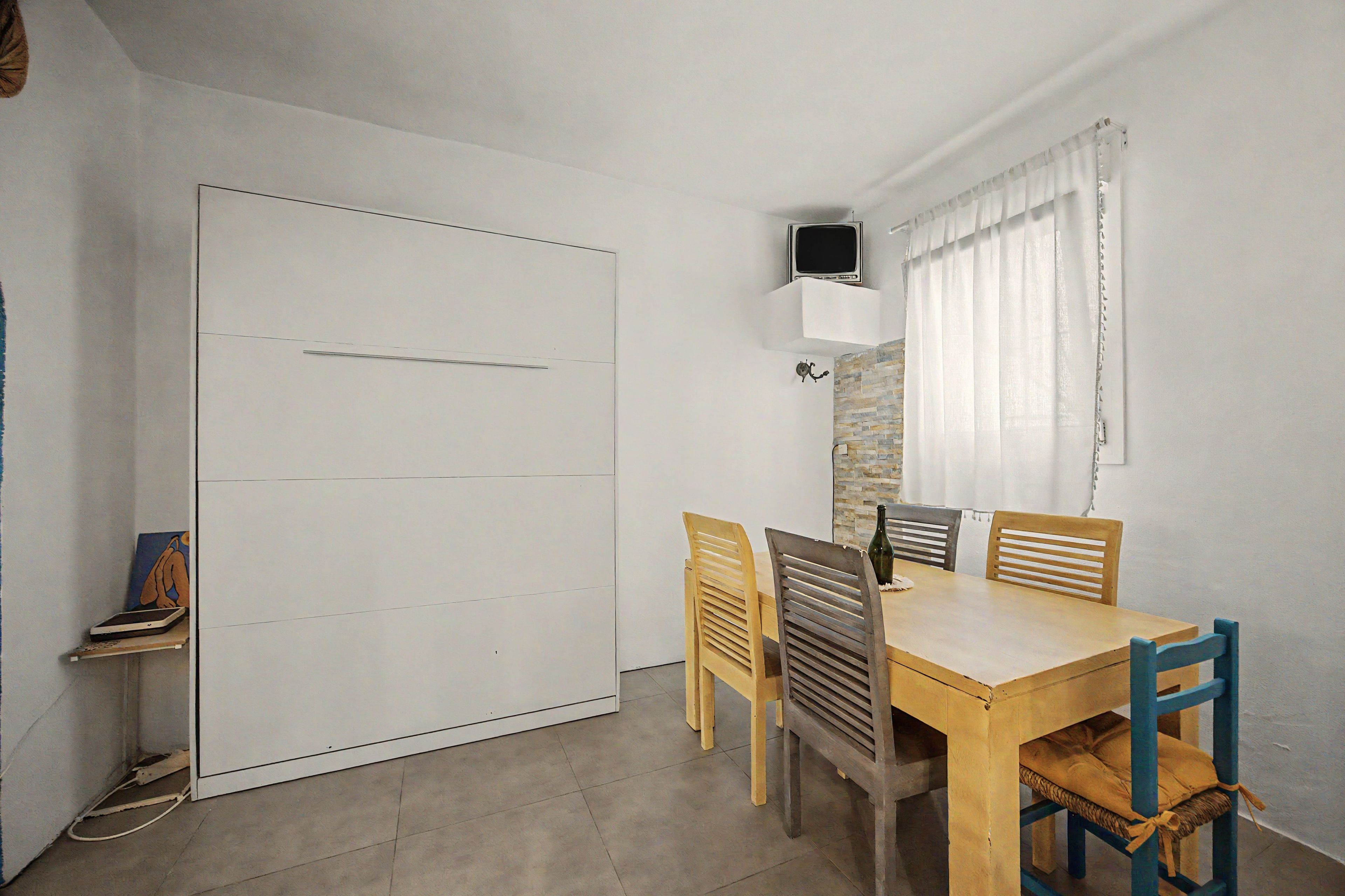 A room with a dining table, four chairs, a television mounted on the wall, and a sliding door.