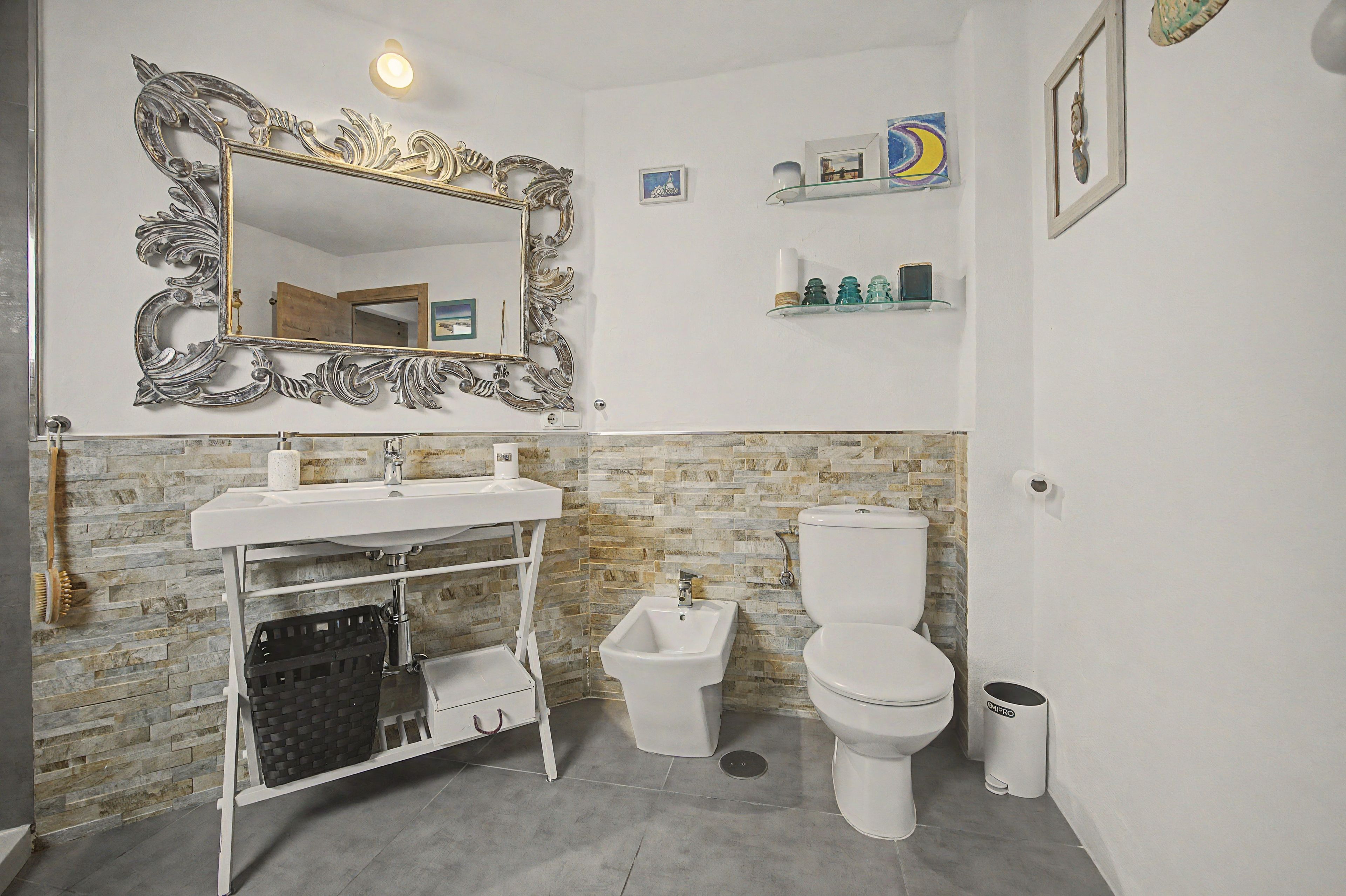 Un baño con pared de piedra, un lavamanos blanco, un inodoro, un espejo con marco decorativo y un pequeño estante con artículos de aseo.