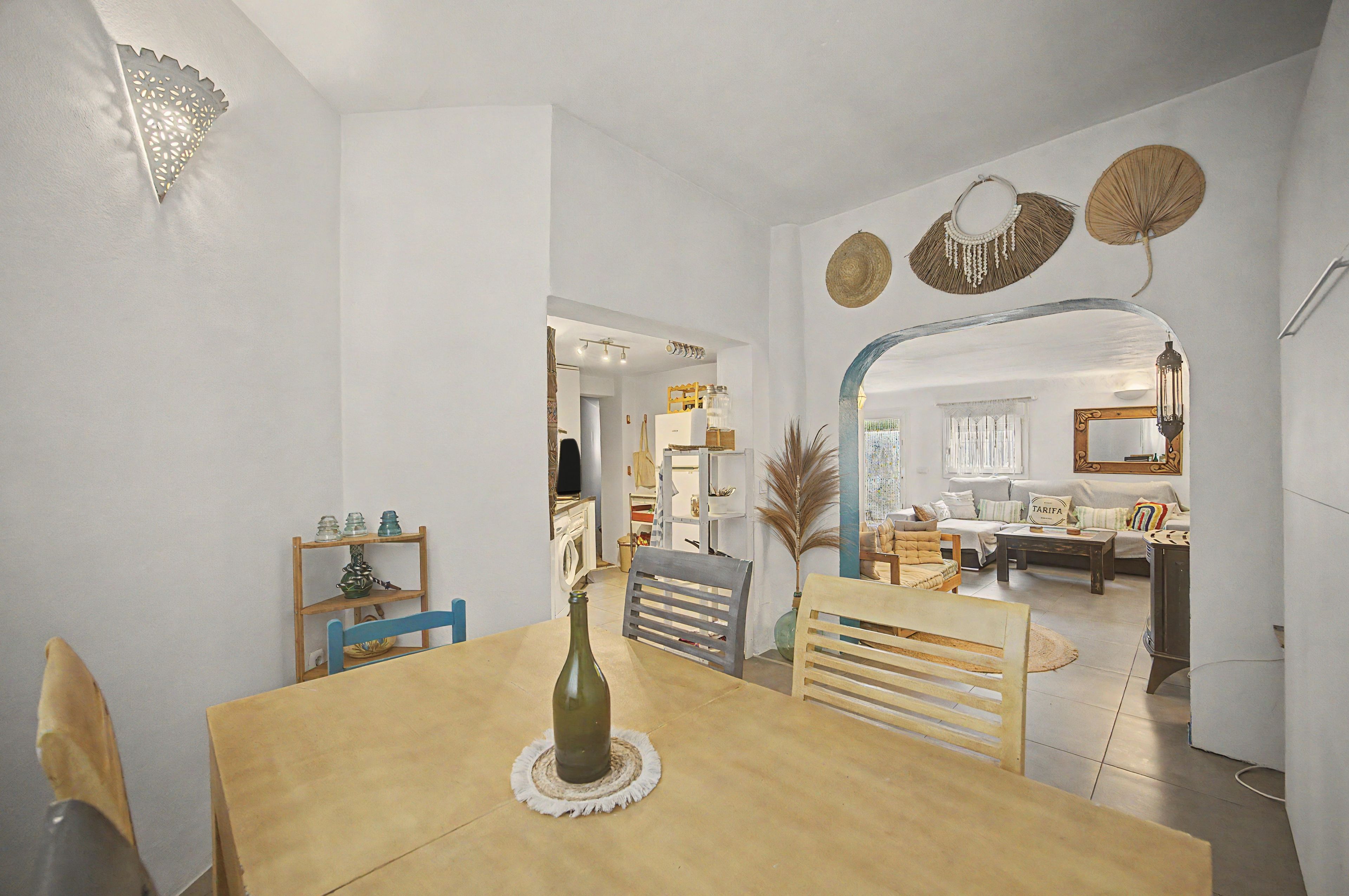 A dining area with a wooden table, chairs, and a bottle of wine. There is a kitchen in the background with a washing machine and cabinets.