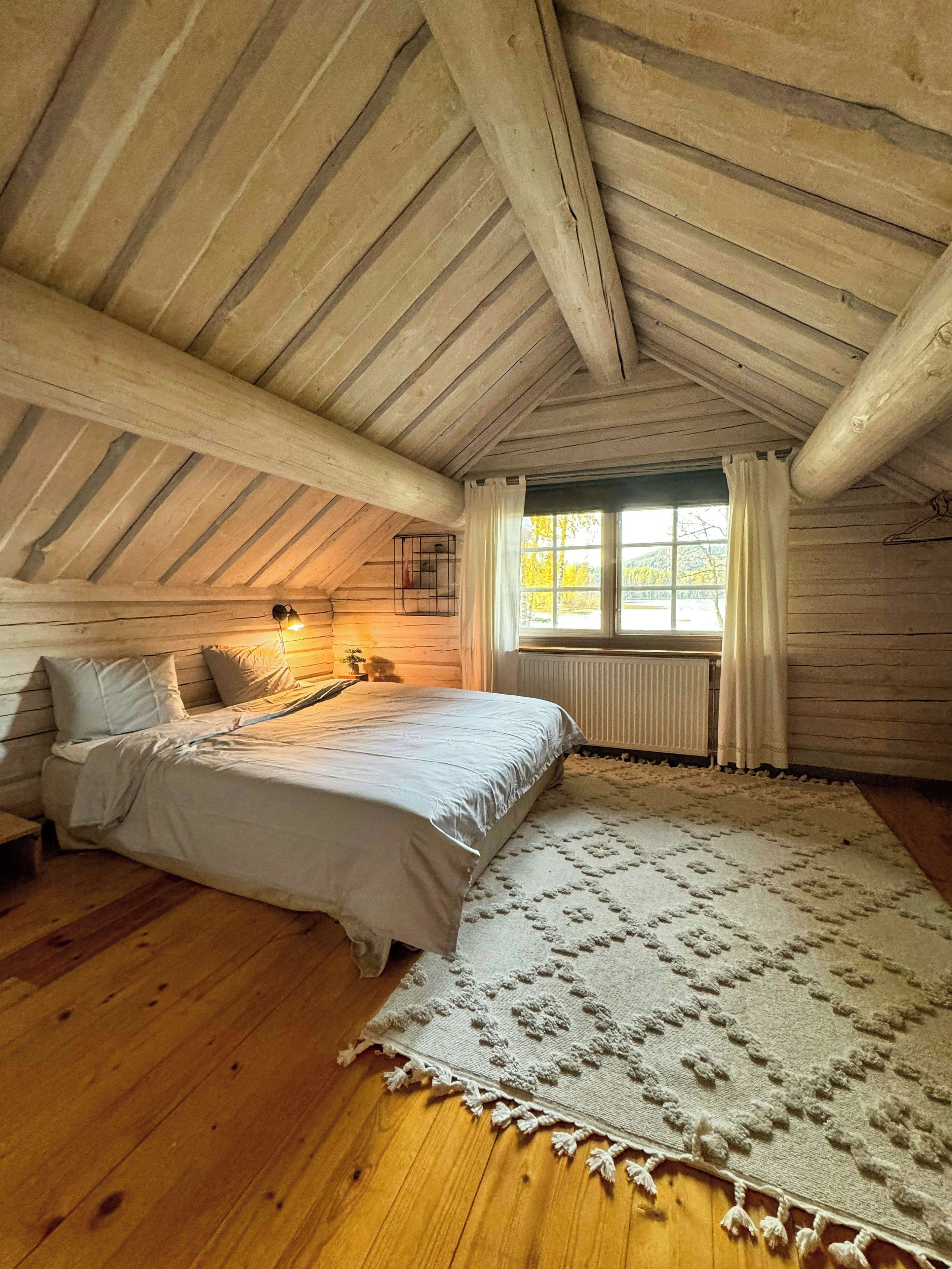 A bedroom with a wooden ceiling, a bed with white bedding, a rug, and a window with curtains.