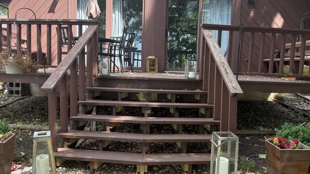 Eine Holztreppe führt zu einer Veranda mit Gartenmöbeln und einem Gartenbereich mit Blumentöpfen.