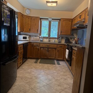 A kitchen with wooden cabinets, a black refrigerator, a microwave, and a window with a view of trees. - New - Lake Ida Cabin (Lake Park)