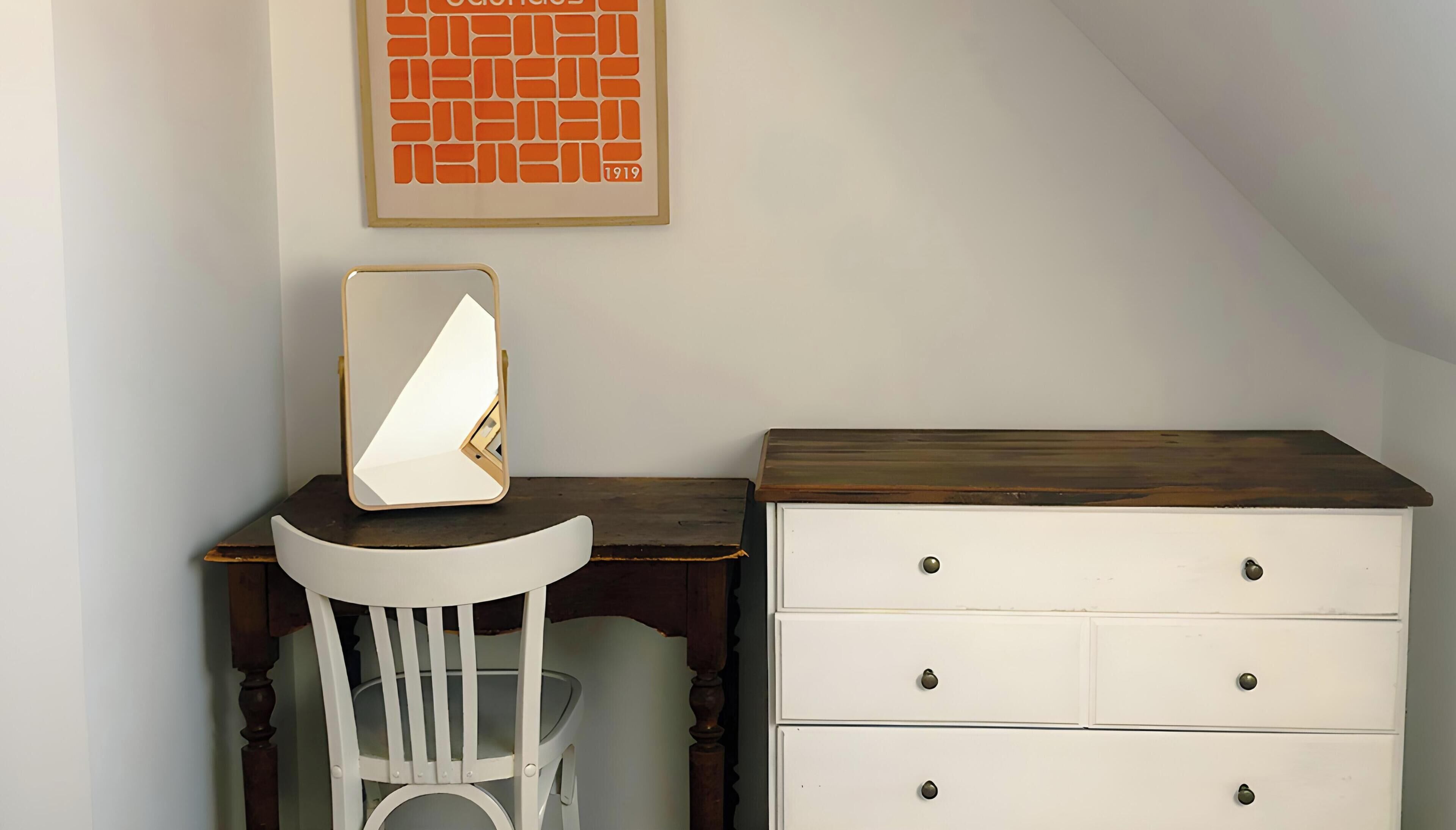 Quarto com cadeira branca, escrivaninha com espelho e cómoda branca com gavetas.