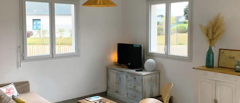 A room with a sofa, a small table, a television on a cabinet, and a window with a view of a garden.