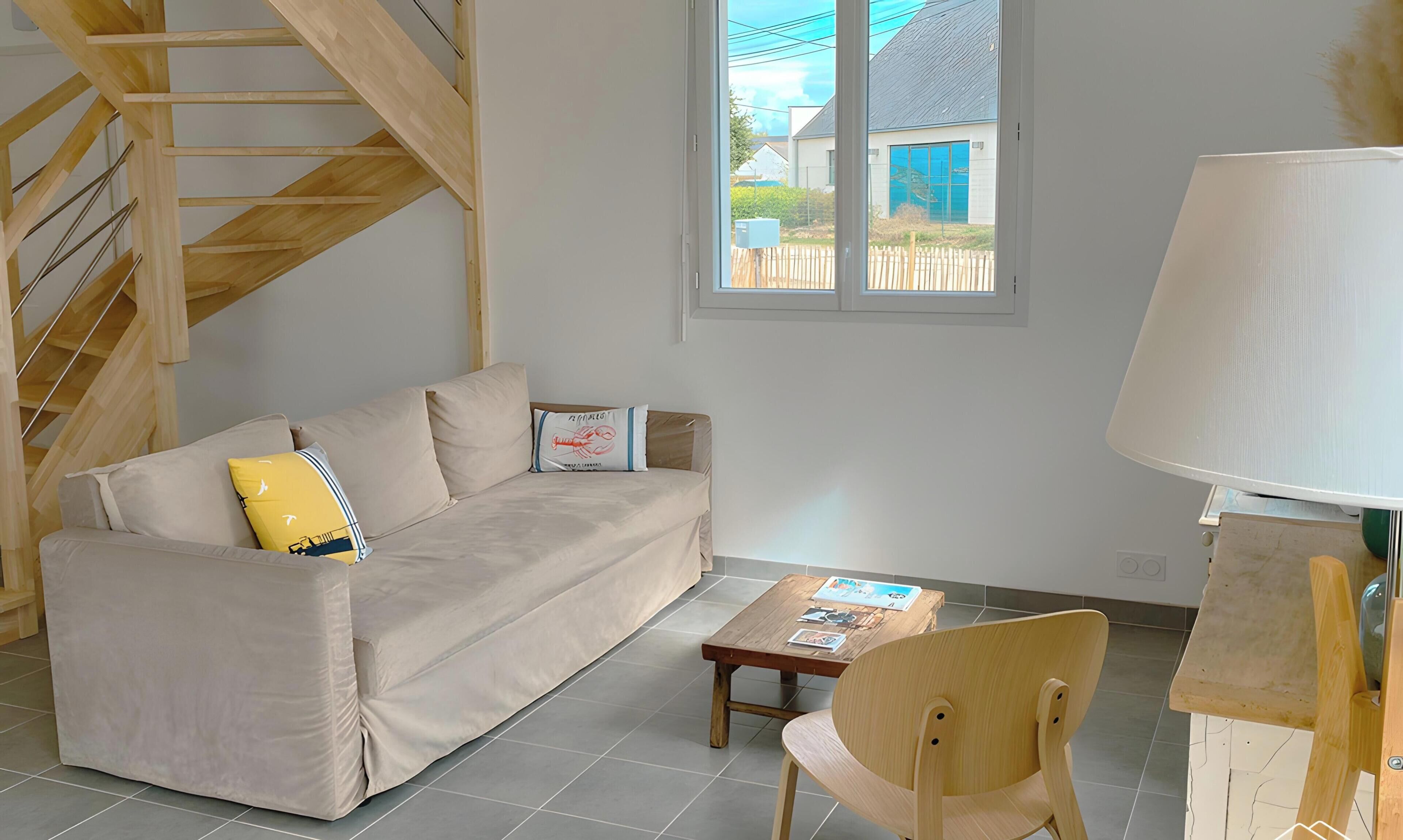 Ein Wohnzimmer mit einer Couch, einem Holzstuhl, einem kleinen Tisch, einer Lampe und einem Fenster mit Blick nach draußen.
