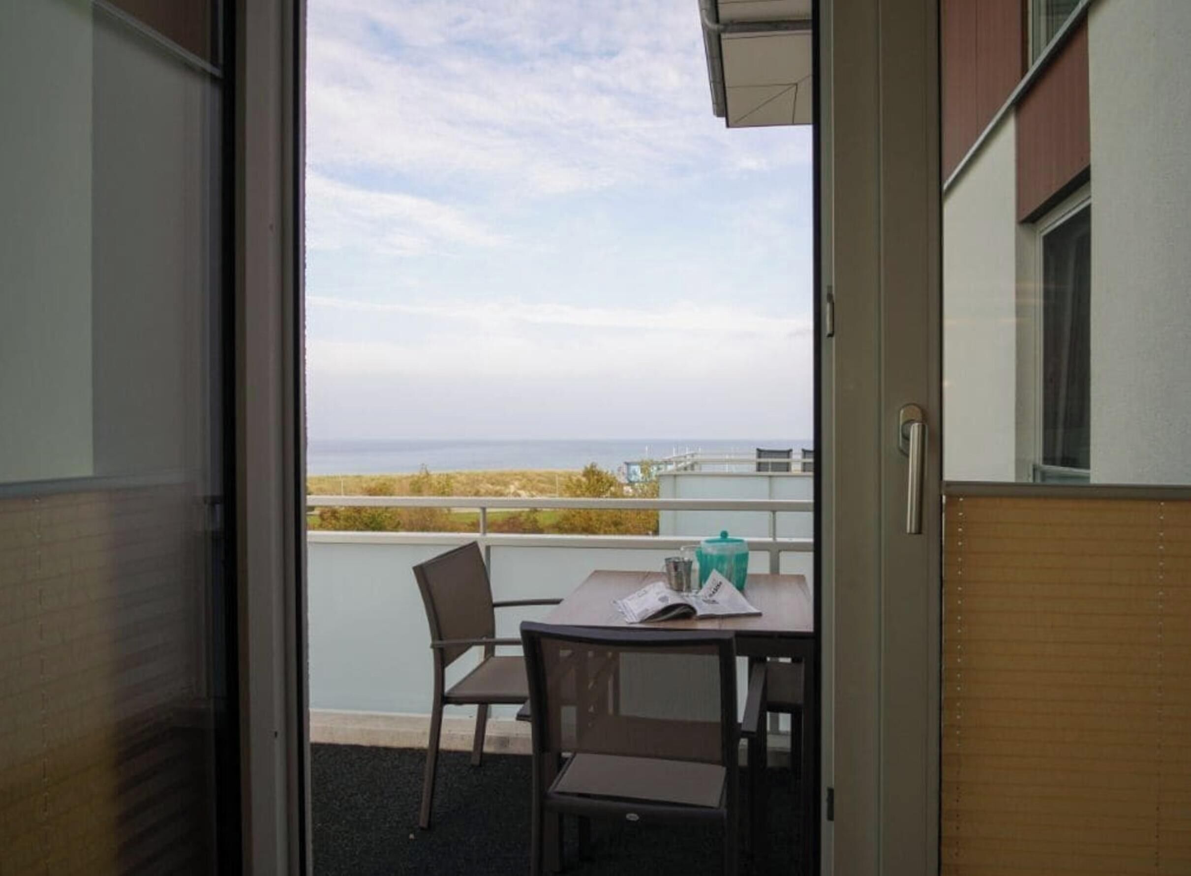 An open door leading to a balcony with a table and chairs, a view of the sea, and a building with a window.