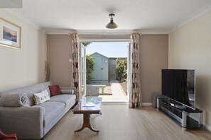 A living room with a sofa, a coffee table, a TV, and a sliding door leading to a garden. - Comfortable Home, Garden & Parking, Walk to Beach! (Westward Ho)
