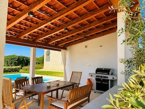 An outdoor patio with a wooden table and chairs, a barbecue grill, and a view of a swimming pool and greenery. - Holiday House with Pool and Olive Trees (Galižana)