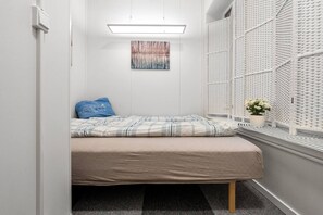 Un dormitorio pequeño y ordenado con una cama, una obra de arte en la pared, una ventana con una celosía blanca y una planta en maceta.