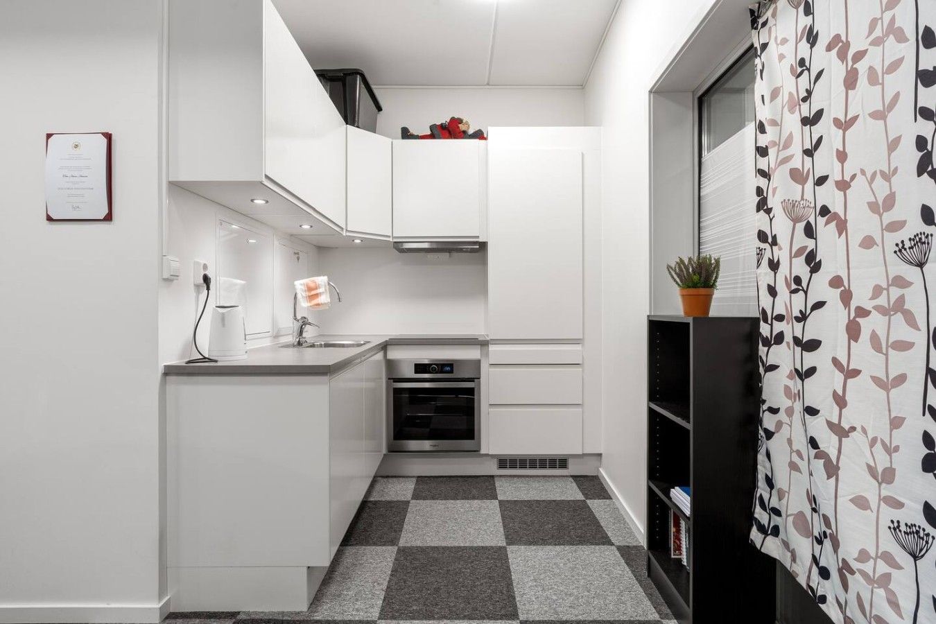 Una cocina compacta con muebles blancos, horno empotrado y suelo de cuadros.