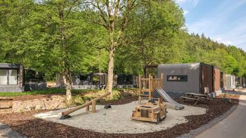 Un parque infantil con tobogán y estructuras para escalar, rodeado de árboles y una zona de grava.