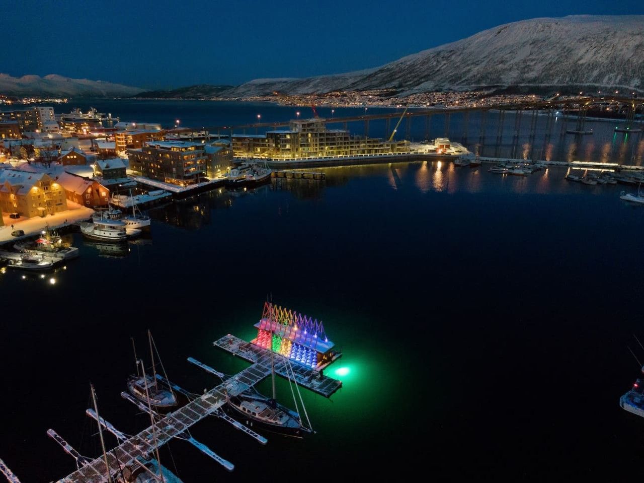 Marina frente al agua de noche, con barcos amarrados, una estructura iluminada y colorida, y un paisaje urbano distante.
