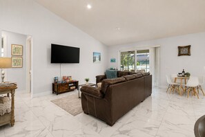 A modern living room with a brown leather sofa, a flat-screen TV mounted on the wall, a small side table with a lamp, and a dining area with a table and chairs. - Cape Coral home with Pool, Spa & Game Room (Cape Coral)