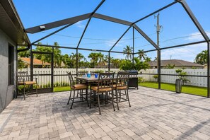 An outdoor patio with a brick floor, a table with chairs, and a grill. - Cape Coral Home with Pool, Spa & Playful Beach House (Cape Coral)