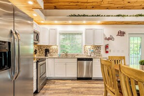 A modern kitchen with white cabinets, stainless steel appliances, and a wooden dining table. - 5 King Bedroom Mtn Lodge Hot Tub, Yard, & Games (Sevierville)