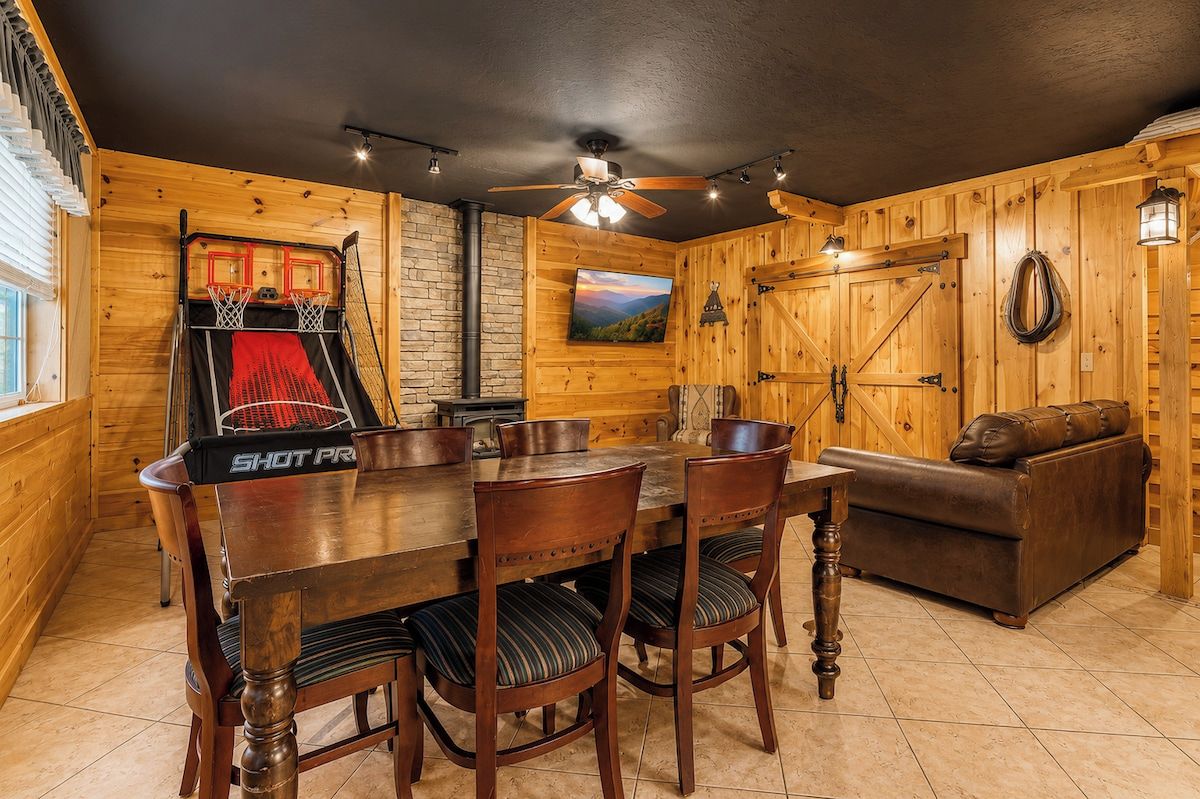 A wooden interior with a dining table, chairs, a sofa, a basketball hoop, and a ceiling fan.