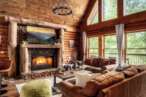 A log cabin interior with a stone fireplace, wooden furniture, and large windows. - Family Retreat! Private Pool, Hot Tub, Game Room! (Sevierville)