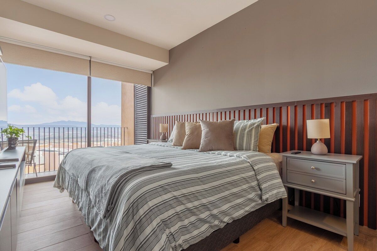 A bedroom with a large window, a bed with striped bedding, a bedside table with a lamp, and a view of the outdoors.