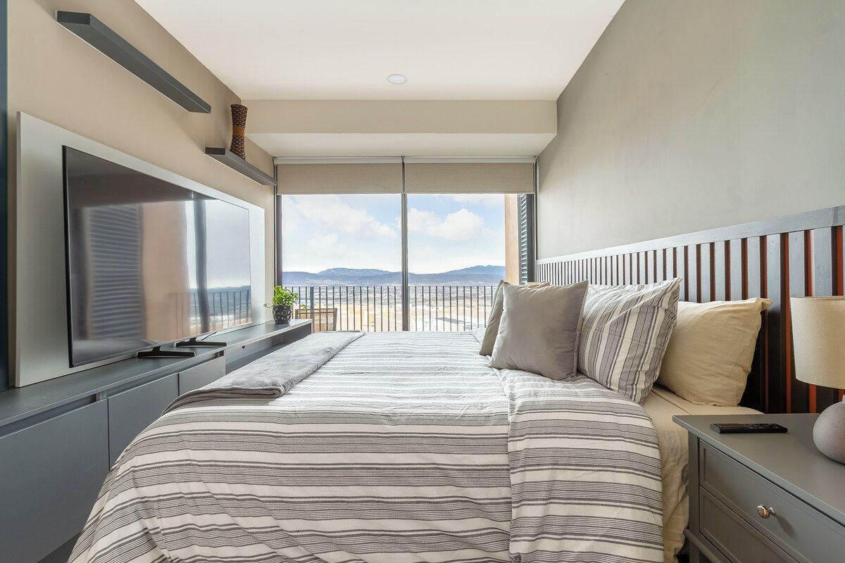 A modern bedroom with a large bed, a TV, and a view of the outdoors.
