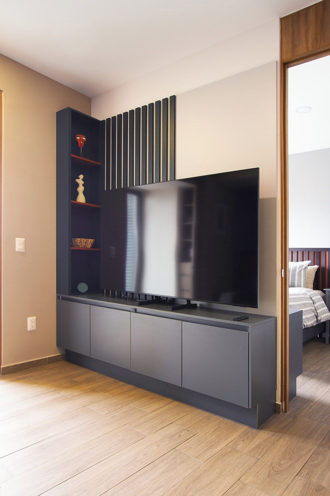 Sala moderna com televisão de tela plana presa a um móvel cinza escuro, piso de madeira e uma cama com roupa de cama listrada ao fundo.