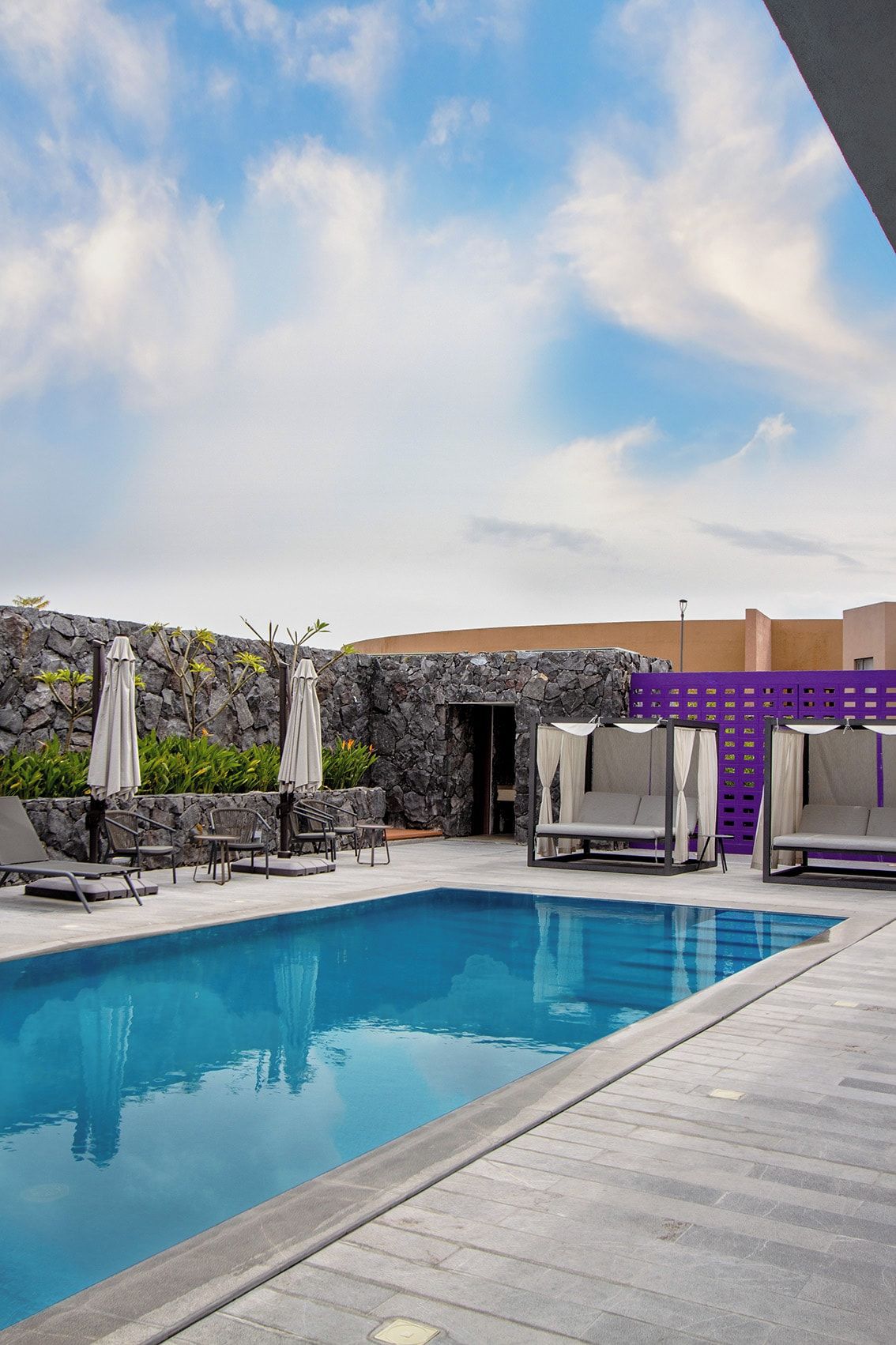 A swimming pool with lounge chairs and umbrellas, surrounded by a stone wall and a purple fence.