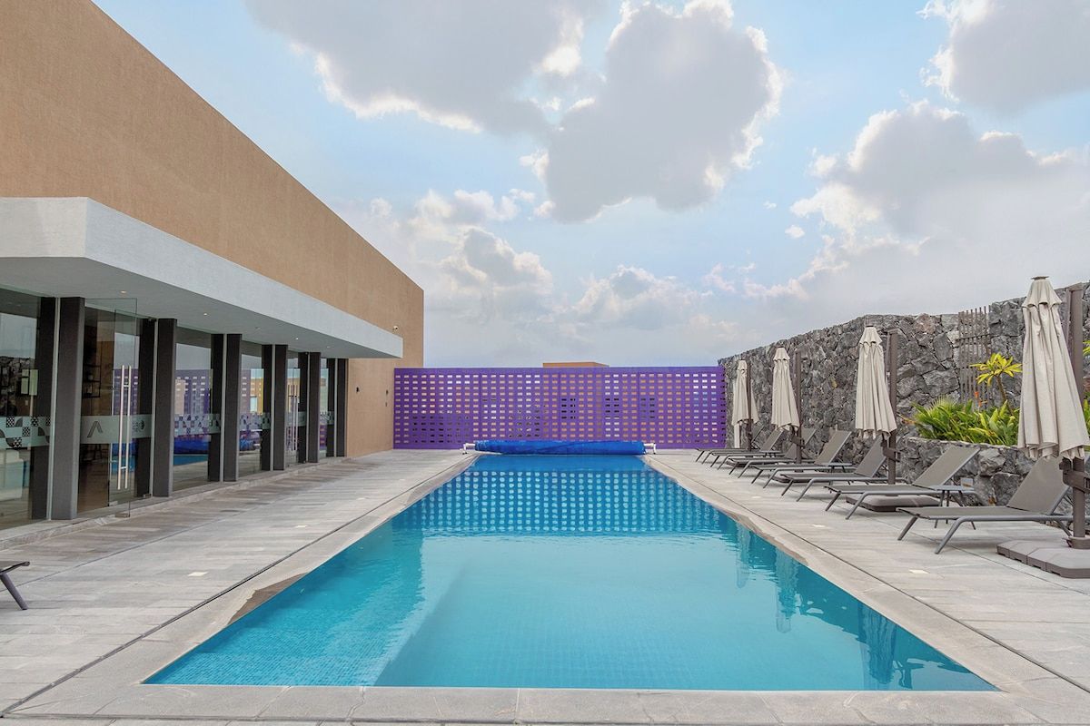 Una piscina con sillas de descanso y sombrillas, rodeada por un edificio con grandes ventanales y un muro de piedra.