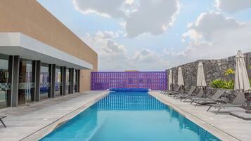 Una piscina con sillas de descanso y sombrillas, rodeada por un edificio con grandes ventanales y un muro de piedra.