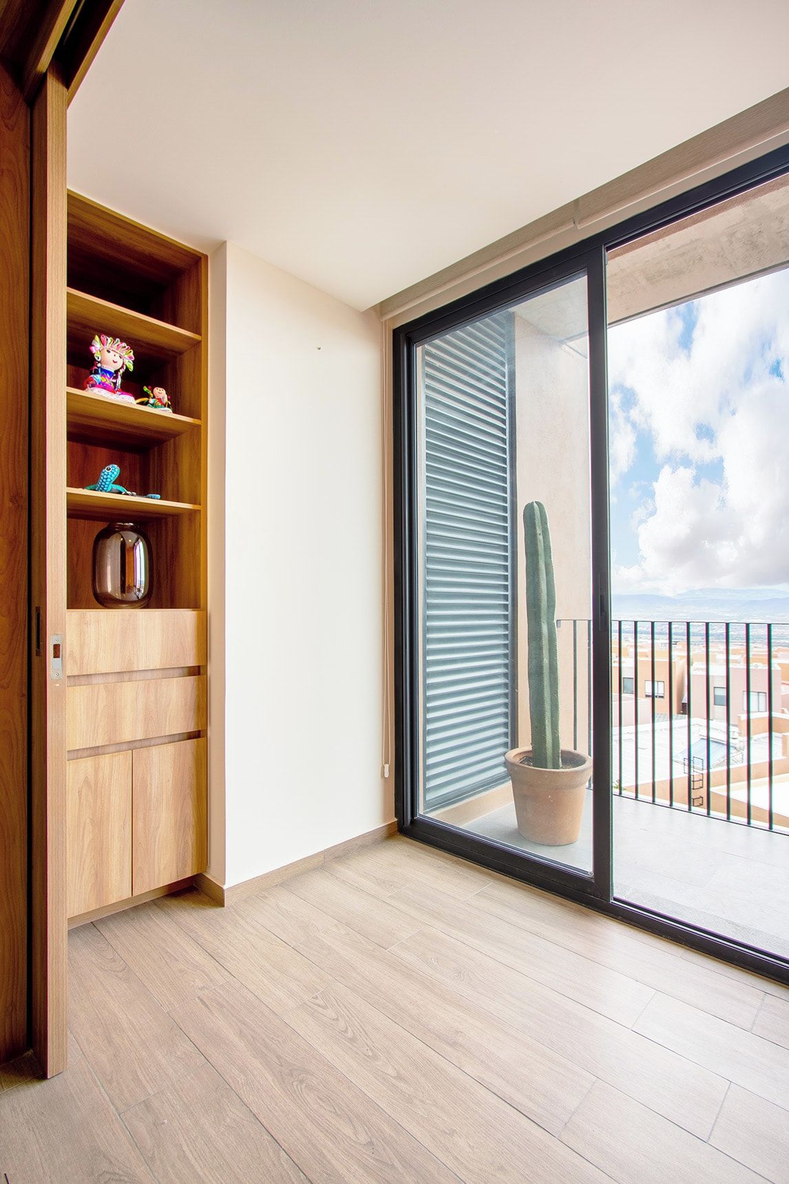 Quarto com armário de madeira, porta de vidro deslizante para uma varanda e um cacto em vaso.