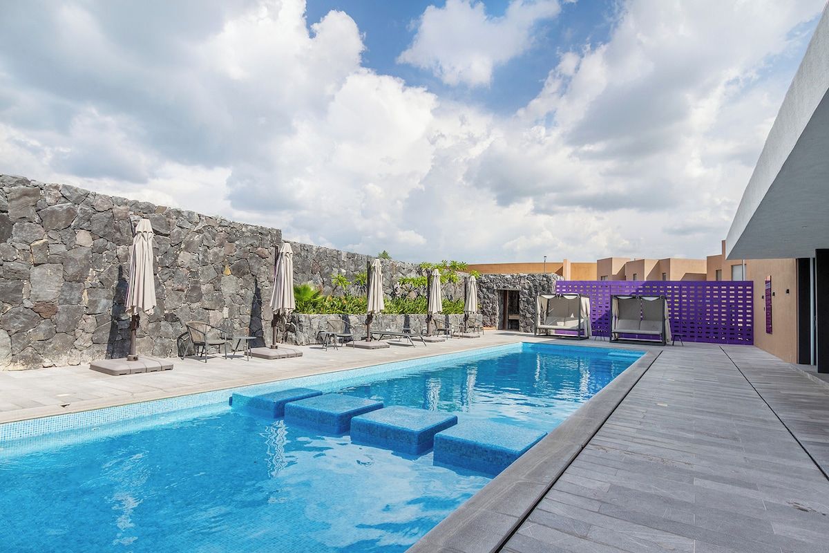 A swimming pool with steps leading into it, surrounded by lounge chairs and umbrellas, with a stone wall and a modern building in the background.